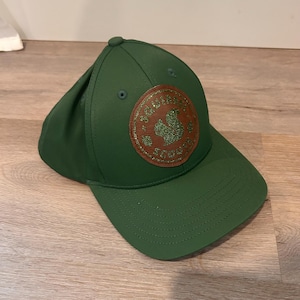 May include: A forest green baseball cap featuring a circular brown leather patch on the front. The patch displays a green glitter squirrel silhouette and the text SQUIRREL SCOUTS embossed in a circular pattern around the central graphic.