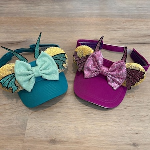 May include: Two visors with a dragon design and a bow. One visor is teal with a light green bow and the other is purple with a pink bow.