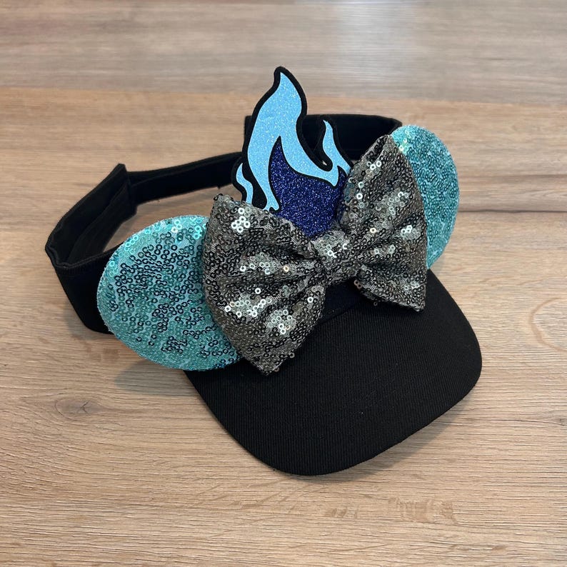 May include: A black visor with a blue and purple flame design, a silver sequined bow, and turquoise sequined ears.