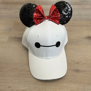 May include: White baseball cap with a black and white Baymax design and a red bow with black sequin mouse ears.