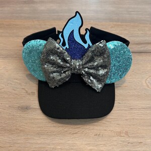 May include: Black baseball cap with a blue and silver sequined bow and a blue flame on top. The cap has turquoise sequined ears on the sides.
