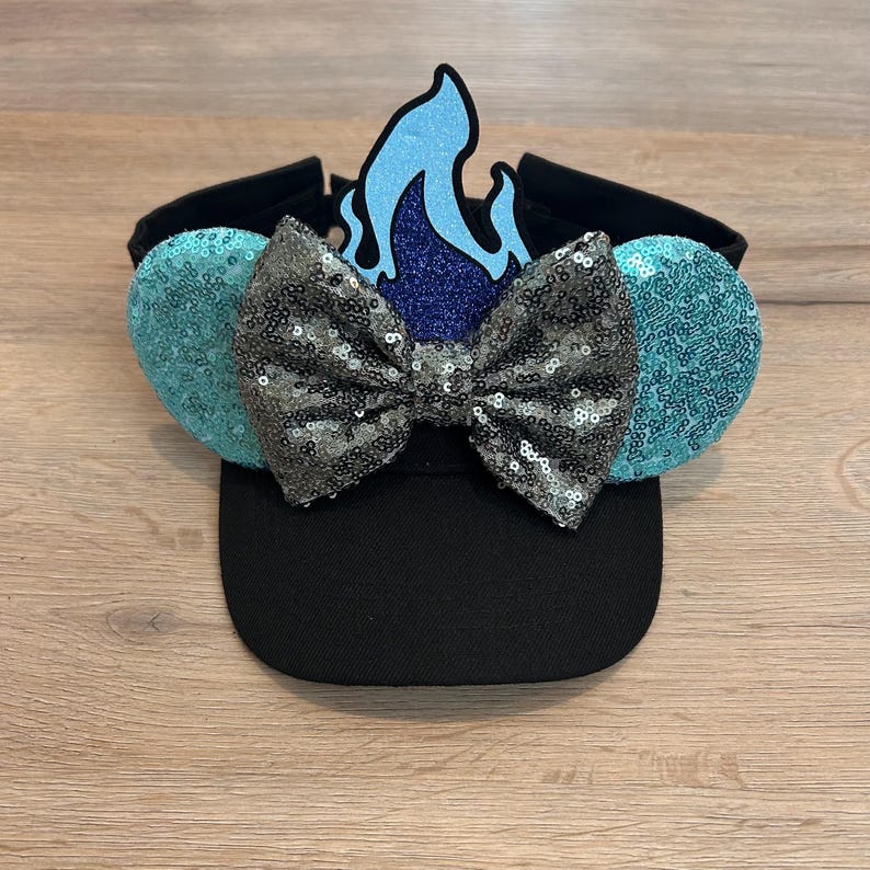 May include: Black baseball cap with a blue and silver sequined bow and a blue flame on top. The cap has turquoise sequined ears on the sides.