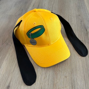 May include: Yellow baseball cap with black ears and a green and white dog collar with a tag that says "Pluto".