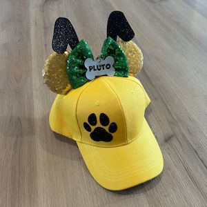 May include: Yellow baseball cap with a black paw print and a green and gold bow with a dog ear headband. The headband has a name tag that says "Pluto".