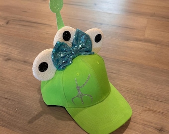 May include: Green baseball cap with a green alien character on the front and white ears with black eyes on the top. The ears have a blue and green bow on top. The alien character has a silver glitter outline.