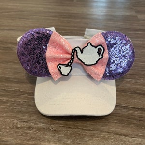 May include: A white baseball cap with a purple and pink bow on top. The bow features a white teapot and teacup with a chain connecting them.