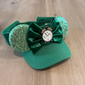 May include: Green visor with a green bow and a gold alarm clock. The clock face has black numbers and hands.