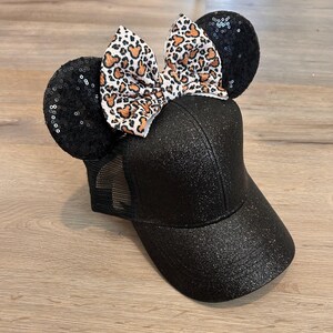 May include: Black glitter baseball cap with black sequin mouse ears and a leopard print bow. The cap has a mesh back and a curved brim. The bow features a white background with brown and orange spots.