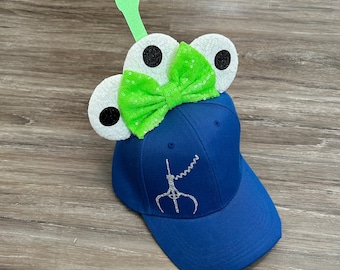 May include: A blue baseball cap with a green bow and white eyes with black pupils. The cap has a silver glitter design of a claw on the front.