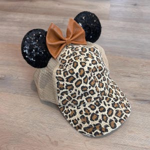 May include: A tan trucker hat with a leopard print front panel and a brown leather bow. The hat features black sequined mouse ears, creating a playful and stylish accessory.