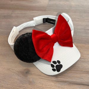 May include: White visor with a black sequin and mesh bow, a red velvet bow, and a black paw print.