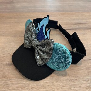 May include: Black visor with a blue and silver sequined bow and a blue flame design. The bow is attached to the visor with a black elastic band.