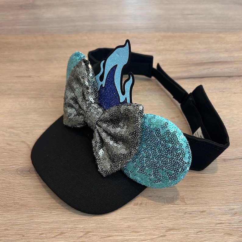 May include: Black visor with a blue and silver sequined bow and a blue flame design. The bow is attached to the visor with a black elastic band.