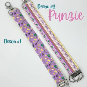 May include: Two purple and yellow patterned keychains with silver key rings. The keychain on the left has a design of a tower, a sun, a chameleon, and a feather. The keychain on the right has a striped pattern. The text "Design #1" is below the left keychain and "Design #2" is above the right keychain. The text "Punzie" is below "Design #2".