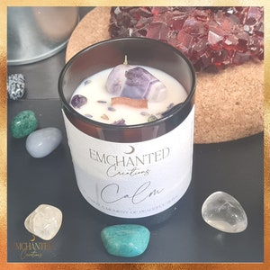 Könnte beinhalten: Ein braunes Glasgefäß mit einer Kerze, auf dem ein weißes Etikett mit der Aufschrift "Enchanted Creations Calm Enjoy a Moment of Peaceful Bliss" angebracht ist. Die Kerze hat einen violetten Amethystkristall obenauf und ist von kleinen Steinen umgeben.