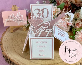 May include: A 3D pop-up greeting card in white and dusty rose featuring a champagne bottle, a glass, and a gift box. The card displays the number 30 and the text: Happy 30th Birthday to our beautiful daughter Lucy. It is a square box-style card.