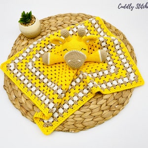May include: A yellow and brown crocheted baby blanket with a crocheted giraffe toy attached. The blanket has a granny square pattern and the giraffe has a white face with black eyes and a black nose.