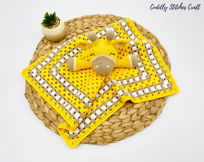 May include: A yellow and brown crocheted baby blanket with a crocheted giraffe toy attached. The blanket has a granny square pattern and the giraffe has a white face with black eyes and a black nose.