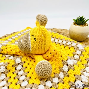 May include: A yellow and brown crocheted baby blanket with a yellow crocheted giraffe toy. The blanket has a checkered pattern and the giraffe has a closed eye.