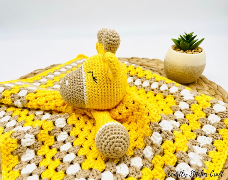 May include: A yellow and brown crocheted baby blanket with a yellow crocheted giraffe toy. The blanket has a checkered pattern and the giraffe has a closed eye.