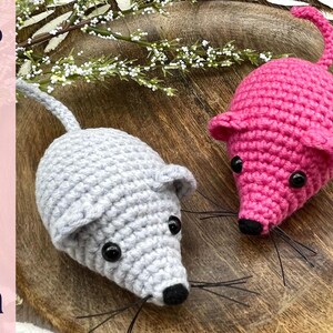 May include: Two crocheted mice, one light gray and one bright pink, are displayed on a wooden surface. Each mouse has black eyes, a black nose, and whiskers. The word "Pattern" is vertically printed on the left side.