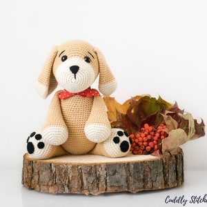 May include: A crocheted beige dog toy with black eyes, a black nose, and black paw prints. The dog is wearing a red bow tie with white polka dots and is sitting on a wooden slice with autumn leaves and red berries.