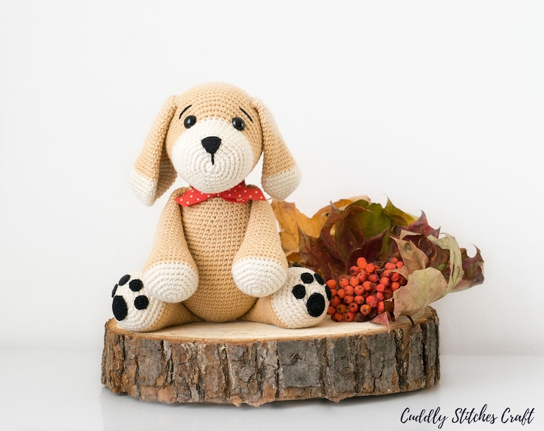 May include: A crocheted beige dog toy with black eyes, a black nose, and black paw prints. The dog is wearing a red bow tie with white polka dots and is sitting on a wooden slice with autumn leaves and red berries.