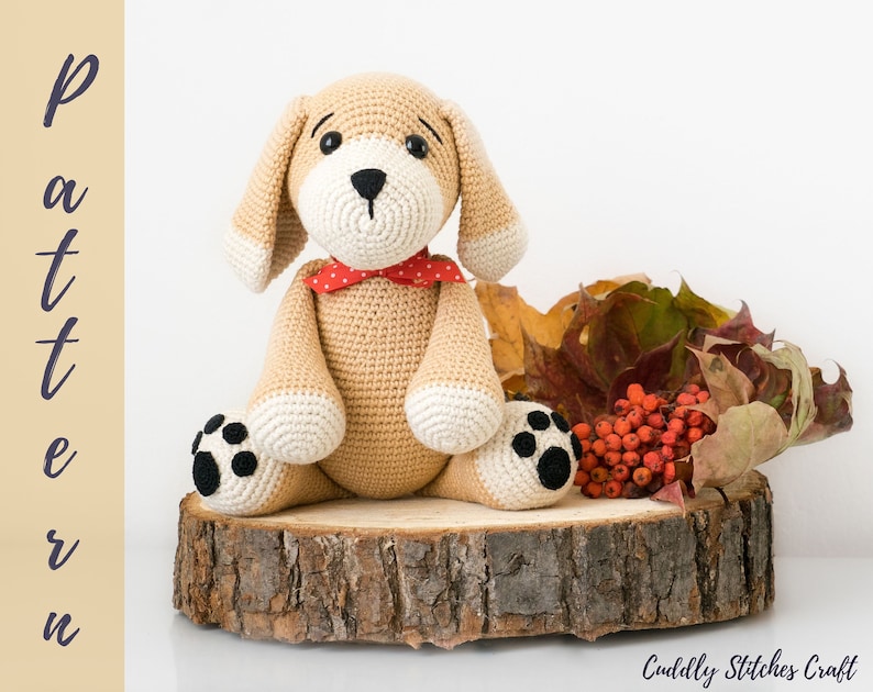 May include: A crocheted stuffed dog toy with brown fur, white paws, and black eyes. The dog is wearing a red bow tie and is sitting on a wooden slice with autumn leaves and red berries.