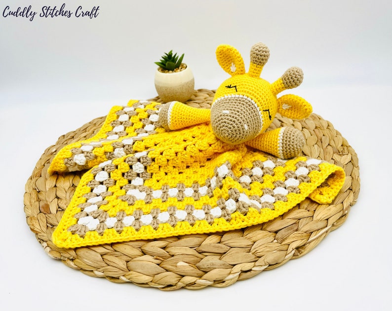 May include: A yellow and brown crocheted baby blanket with a crocheted giraffe toy on top. The blanket has a checkered pattern and the giraffe has closed eyes.