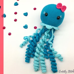 Puede incluir: Un juguete de amigurumi de medusa tejido a crochet con un cuerpo azul y turquesa, un lazo rosa y ojos negros. El juguete está sobre un fondo blanco con corazones azules y rosas esparcidos.