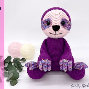 Puede incluir: Juguete de amigurumi de perezoso de crochet morado con cara rosa y ojos coloridos. El perezoso está sentado con los brazos y las piernas extendidos.