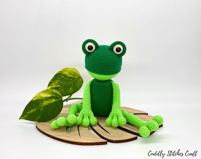 May include: A green crocheted frog sitting on a wooden leaf-shaped base. The frog has black eyes and a light green belly.