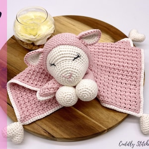 May include: A pink crocheted baby blanket with a white crocheted lamb attached. The blanket has a heart-shaped tag on each corner. The lamb has closed eyes and a pink nose.