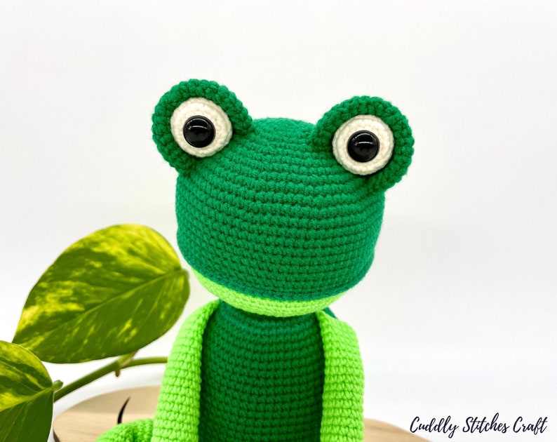 May include: Green crocheted frog amigurumi toy with black button eyes. The frog is sitting on a wooden surface.