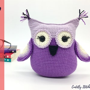 Puede incluir: Juguete de amigurumi de búho de crochet morado con ojos negros y pico negro. El búho tiene detalles de hilo blanco en sus alas y orejas.