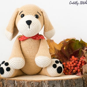 May include: A crocheted beige puppy with black eyes, a black nose, and white paws. The puppy is wearing a red and white polka dot bow tie and is sitting on a wooden log surrounded by fall leaves and red berries.
