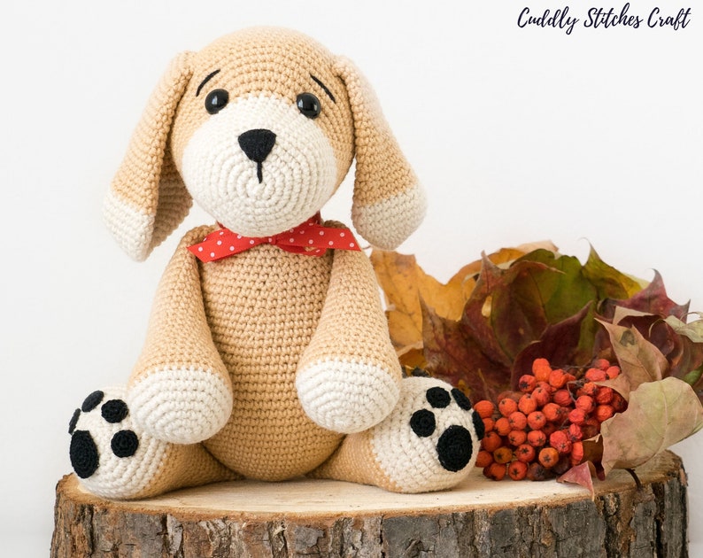 May include: A crocheted beige puppy with black eyes, a black nose, and white paws. The puppy is wearing a red and white polka dot bow tie and is sitting on a wooden log surrounded by fall leaves and red berries.