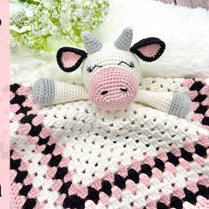 May include: A crocheted white, pink and black baby blanket with a crocheted cow toy on top. The blanket is made with a granny square pattern.