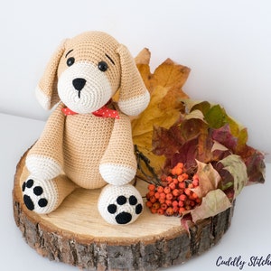 May include: A crocheted beige dog toy with black paw prints, red and white polka dot bow tie, and brown eyes. The toy is sitting on a wooden slice with fall leaves and red berries.