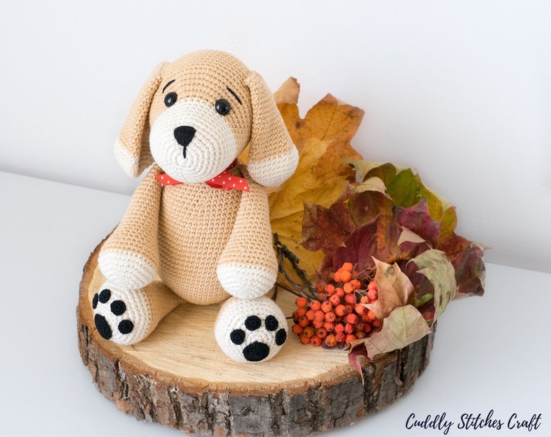 May include: A crocheted beige dog toy with black paw prints, red and white polka dot bow tie, and brown eyes. The toy is sitting on a wooden slice with fall leaves and red berries.