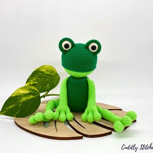 May include: A green crocheted frog sitting on a wooden leaf-shaped base. The frog has large black eyes and a light green belly.