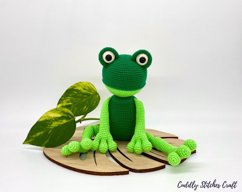 May include: A green crocheted frog sitting on a wooden leaf-shaped base. The frog has large black eyes and a light green belly.
