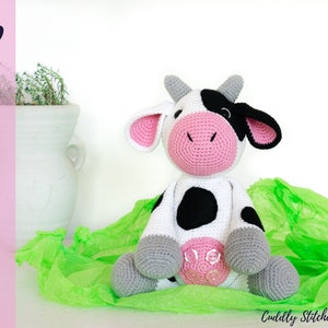 Pode incluir: Um brinquedo de pelúcia de vaca de crochê com manchas pretas e brancas, orelhas cor-de-rosa e um nariz cor-de-rosa. O brinquedo está sentado em uma superfície verde.