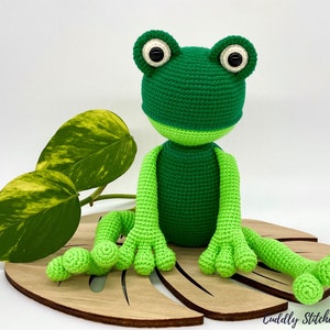 May include: A green crocheted frog sitting on a wooden leaf. The frog has large black eyes and a light green belly.
