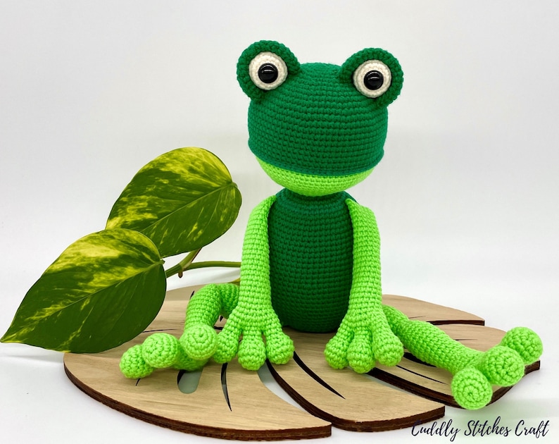 May include: A green crocheted frog sitting on a wooden leaf. The frog has large black eyes and a light green belly.