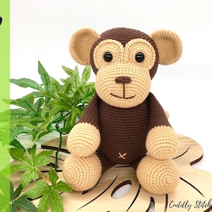 May include: A crocheted monkey toy with brown fur, beige arms and legs, and black eyes. The monkey is sitting on a wooden leaf-shaped base.