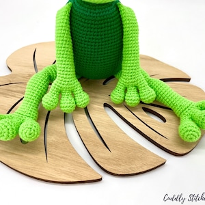 May include: A green crocheted frog sitting on a wooden monstera leaf. The frog has a green body and long green legs with four toes on each foot.