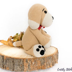 May include: A crocheted beige dog with white accents and a red bow tie sitting on a wooden slice. The dog has black eyes and a black paw print on one foot.