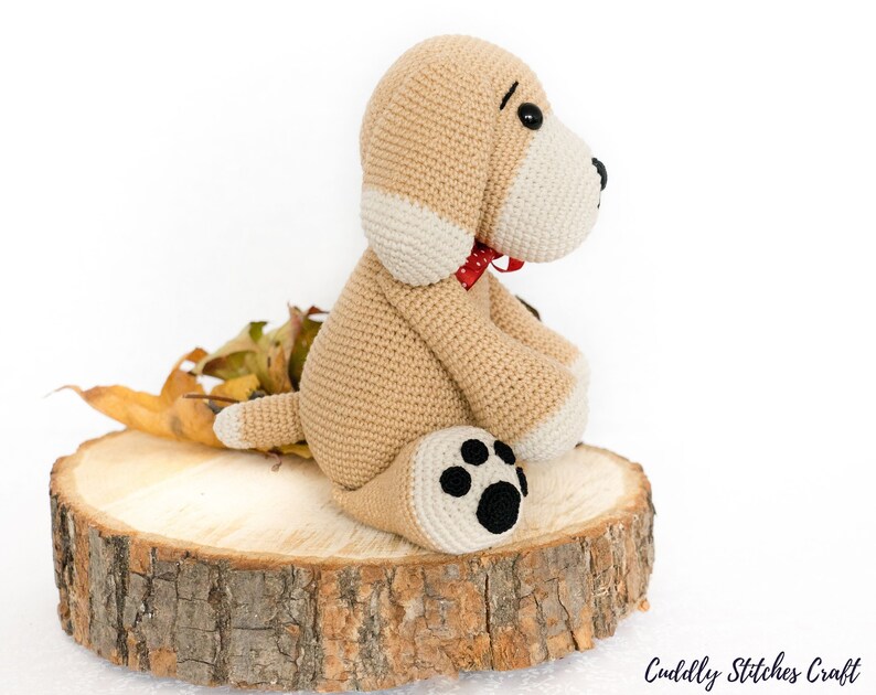 May include: A crocheted beige dog with white accents and a red bow tie sitting on a wooden slice. The dog has black eyes and a black paw print on one foot.