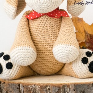 May include: A crocheted beige dog with white paws and black paw pads. The dog is wearing a red and white polka dot bow tie. The dog is sitting on a wooden log.